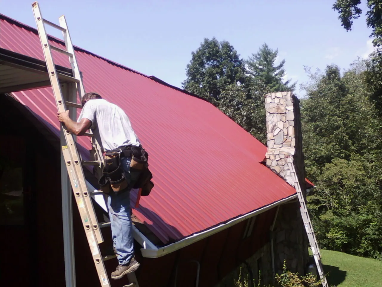 Roofer on a ladder beside a red metal roof and stone chimney — Soffit & Fascia Repair by Roofing Brothers in Epworth